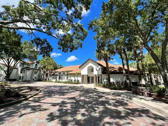 Inviting apartment complex entrance with charming architecture, surrounded by lush greenery and a picturesque cobblestone pathway under a bright blue sky.