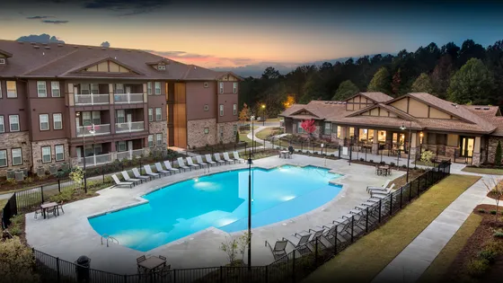 Luxurious apartment complex with modern architecture, featuring a sparkling swimming pool surrounded by stylish lounge chairs and lush greenery at sunset.