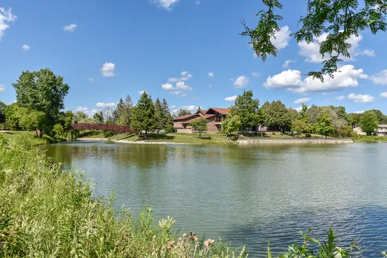 Picturesque view of a serene lake and charming residential area enhanced by lush greenery and a quaint red bridge under a bright blue sky.