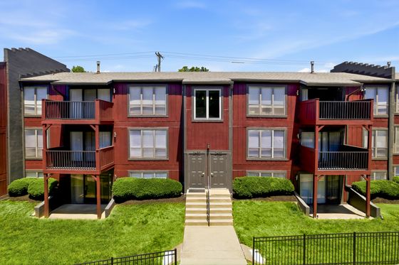 Charming red apartment building with balconies, surrounded by lush greenery, offering a welcoming entrance and bright blue sky overhead.