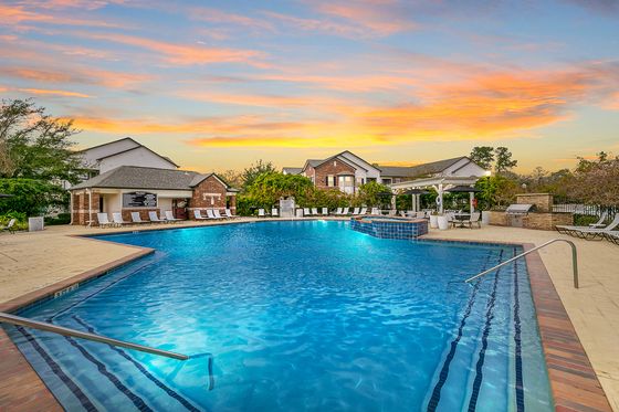Luxurious apartment complex with a serene pool, sunlit skies, and inviting seating areas, perfect for relaxation.