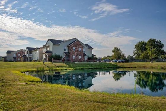 Charming residential complex with picturesque pond, lush green landscape, and inviting two-story brick-and-siding homes under a bright blue sky.