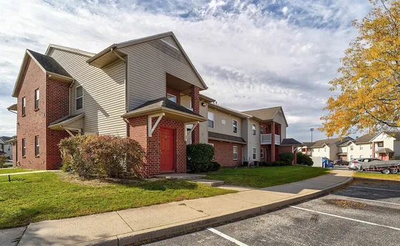 Charming residential complex with red brick accents and a lush grassy area, offering ample parking and vibrant autumn foliage under a bright sky.