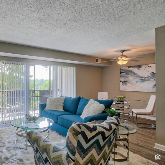 Inviting living room with a stylish blue sofa, modern glass tables, and chevron-patterned armchair, opening onto a bright balcony view.