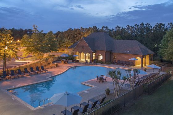 Serene evening view of a luxury apartment complex featuring a large illuminated swimming pool, comfortable lounge chairs, and inviting clubhouse surrounded by lush greenery.