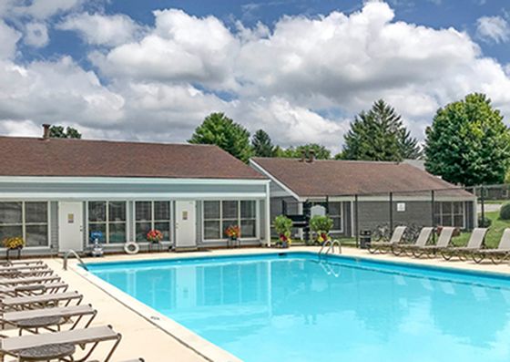 Inviting outdoor pool area with sun loungers and a modern clubhouse, surrounded by lush greenery and a bright blue sky.