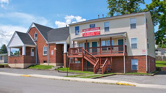 Sunny street view of charming brick and siding apartment building with inviting front porch, surrounded by lush greenery