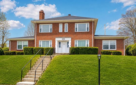 Charming red-brick two-story house with white accents, nestled on a manicured green lawn under a clear blue sky.