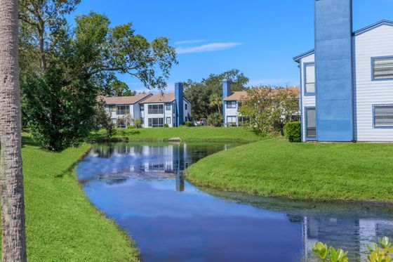 Tranquil apartment view featuring charming blue and white buildings, vibrant greenery, and a serene pond under a clear blue sky.
