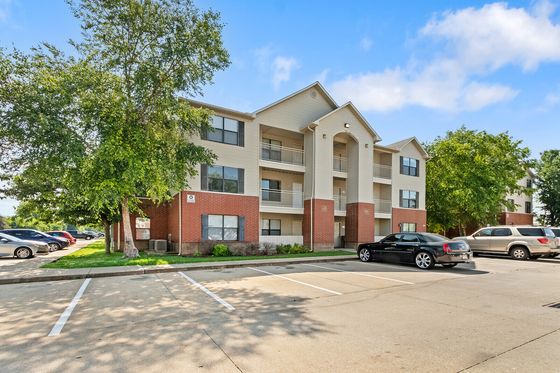 Charming three-story apartment building with modern architecture, surrounded by lush greenery and ample parking space on a sunny day.