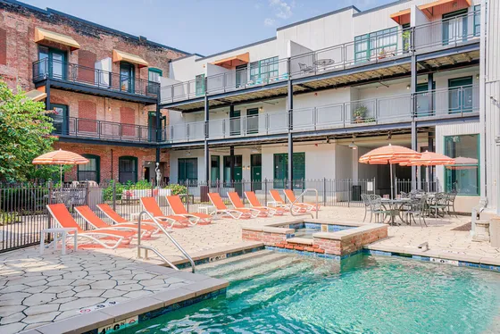 Charming apartment complex with a vibrant courtyard, featuring a refreshing pool, sun loungers, and patio seating under cheerful striped umbrellas.