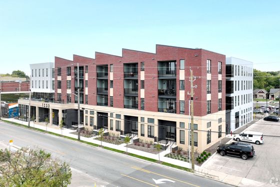 Modern apartment building with red brick exterior, balconies, and ample parking in an urban setting.