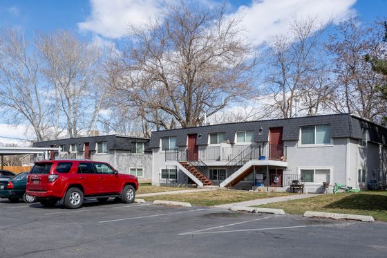 Charming two-story apartment complex with well-maintained parking area, featuring vibrant red doors and surrounded by lush trees under a clear blue sky.