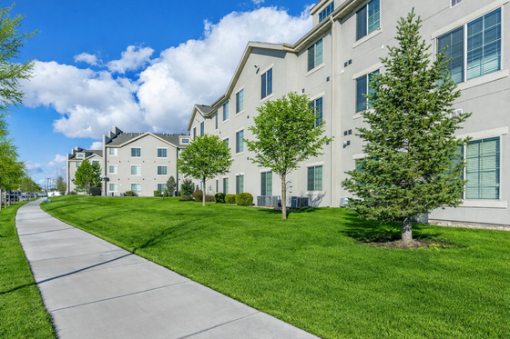 Charming apartment building with lush green landscaping and a winding pathway, set against a clear blue sky.