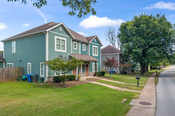 Charming two-story green house with white trim on a serene residential street, featuring a well-maintained lawn and inviting front porch.