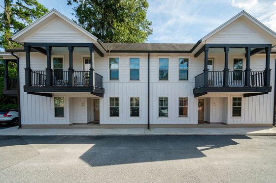 Charming two-story apartment building with elegant black railing balconies and crisp white siding, set against a lush green backdrop.