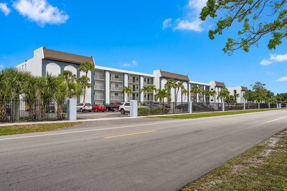 Modern apartment complex with tropical landscaping, ample parking, and sleek architectural design under a clear blue sky.