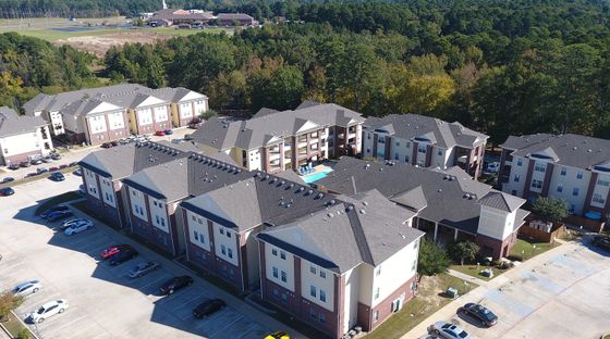 Aerial view of modern apartment complex with ample parking and surrounded by lush greenery, showcasing a serene and spacious living environment.