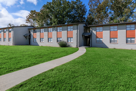 Charming two-story apartment building surrounded by lush green grass and shaded by mature trees, offering a serene living environment.