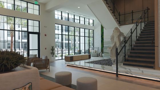 Elegant apartment lobby with tall glass windows, modern wooden furniture, and a sleek staircase, creating a welcoming atmosphere.