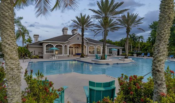 Elegant poolside view of a luxury apartment complex, featuring palm trees, sun loungers, and a clubhouse under a serene evening sky.