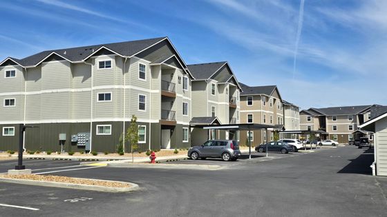 Modern apartment complex with multiple levels, contemporary architecture, and spacious parking area, set against a clear blue sky.
