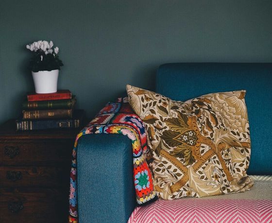 Cozy living room corner with a vibrant blue sofa, adorned with a vintage-style patterned pillow and colorful knit throw, next to a classic wooden side table with a stack of antique books and a potted plant.