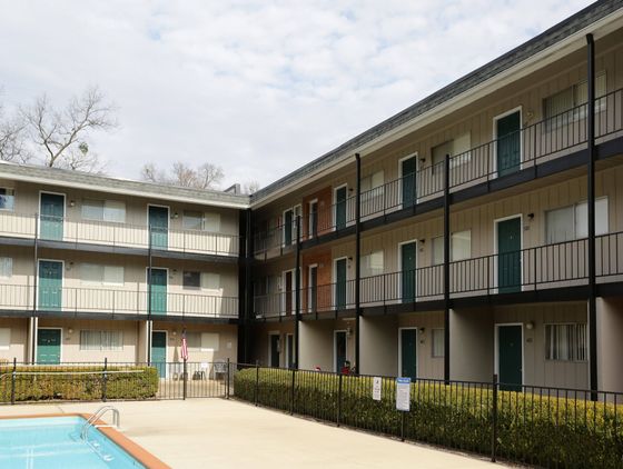 A multi-story apartment building with balconies facing a swimming pool. The building features neutral-colored siding and metal railings. The pool area is enclosed with a fence, and there are lounge chairs and umbrellas around the pool.