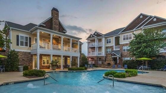 A well-maintained pool area with clear water, surrounded by lush greenery. Two apartment buildings with large windows and a stone facade are visible in the background. The scene is set during dusk, with soft lighting creating a warm atmosphere.