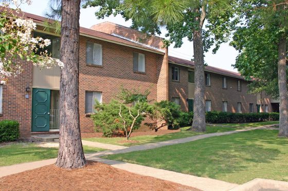 A two-story brick apartment building surrounded by large trees and well-kept green grass. The building has multiple windows, and the landscape features tidy garden beds with shrubs.
