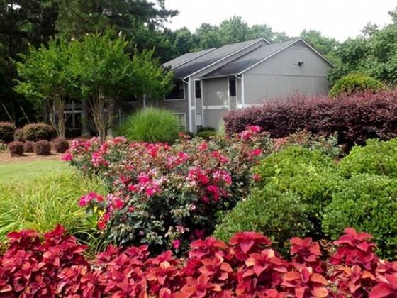 A cozy leasing office surrounded by vibrant flower beds, including pink, purple, and red flowers. The landscaping includes lush green plants, and the building itself is gray with a simple yet welcoming design.