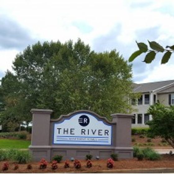 The entrance sign for The River apartment complex, featuring large white and blue lettering with the text 'THE RIVER' on a large stone base. The sign is surrounded by well-maintained landscaping and trees, with the apartment building visible in the background.