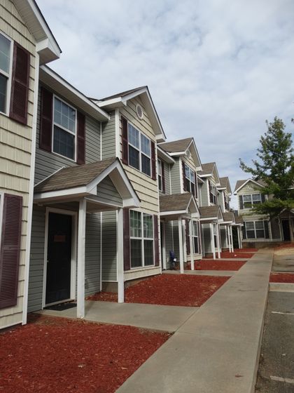 A multi-unit apartment building with a light-colored exterior, featuring several windows and a well-maintained walkway. The area around the building is landscaped with red mulch, trees, and green shrubs.