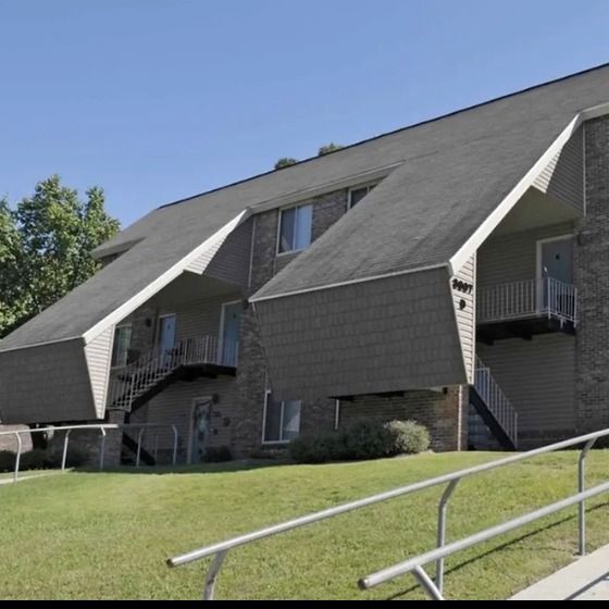 A multi-story apartment building with beige siding and a grassy slope leading to the entrance. The building features several balconies, and the landscape includes well-trimmed bushes and small trees.