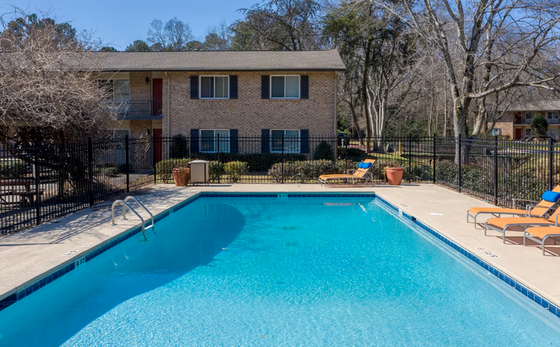 A sparkling blue swimming pool in front of a two-story brick apartment building. The pool area includes lounge chairs, a decorative potted plant, and a green fence surrounding the space.