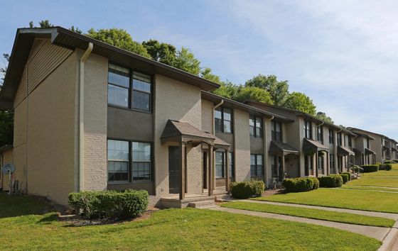 An apartment building with a tan exterior, featuring several windows and a well-maintained walkway leading to the entrance. Green landscaping surrounds the building, with trees and shrubs along the path.