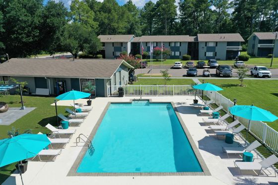 Aerial view of a swimming pool at an apartment complex, surrounded by lounge chairs with turquoise umbrellas. The pool is situated near a clubhouse, with lush green trees and apartments in the background.