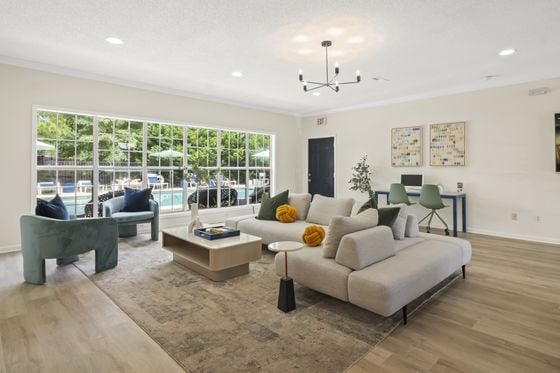 A modern and stylish leasing office or clubhouse living room, featuring large windows with a view of the outdoor pool area. The room is furnished with a light gray sofa, green accent pillows, and a sleek coffee table. In the background, there is a dining area and a stylish chandelier hanging from the ceiling, creating a welcoming atmosphere.