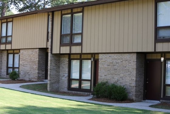 A two-story apartment building with a tan and brown exterior, surrounded by a well-maintained lawn. The landscape includes several trees and bushes, contributing to a green and inviting atmosphere.he building has several windows and a paved sidewalk leading to the entrance, with neatly trimmed hedges along the path.