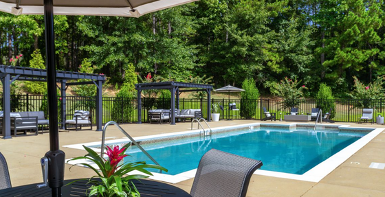 A bright and inviting swimming pool with crystal-clear water, surrounded by lounge chairs and a table with an umbrella. A gazebo with seating is visible in the background, and lush greenery surrounds the area.