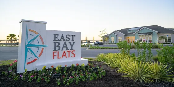 Entrance sign for East Bay Flats apartments, featuring the text 'EAST BAY FLATS' in large letters. The sign is surrounded by well-maintained landscaping with colorful flowers and palm trees. The background shows a modern apartment building with a large parking area and water in the distance.