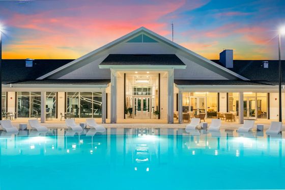 A beautifully lit clubhouse with large windows, overlooking a sparkling swimming pool. The pool area is surrounded by lounge chairs and umbrellas. The sky is illuminated with vibrant colors from a sunset, adding a serene atmosphere to the scene.