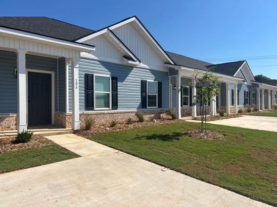 A single-story row of townhome buildings with a light blue exterior and a neat front yard. The building has several windows, and the lawn is well-kept with small shrubs and trees scattered along the landscape.