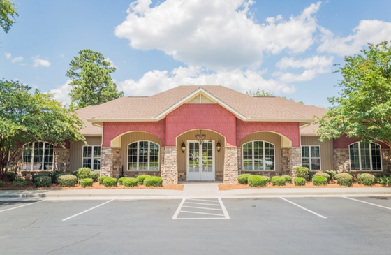 Exterior view of an apartment leasing office with a red brick facade, large windows, and neatly trimmed landscaping. The parking lot in front is paved, with a clear blue sky above.