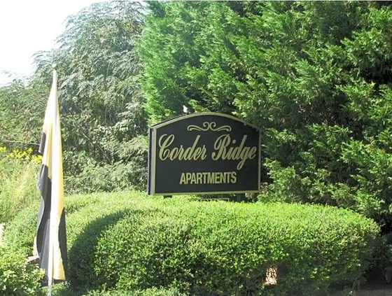 A black and gold sign with the text 'Corder Ridge Apartments' located near a well-maintained hedge and green shrubbery, indicating the entrance to the apartment complex