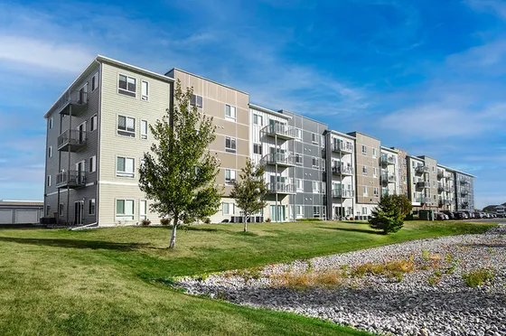 Modern apartment complex with four-story buildings, spacious balconies, and lush green surroundings under a clear blue sky.