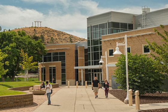 Modern university campus building featuring sleek architecture, large glass windows, and scenic hill views, with students walking on a sunlit path.