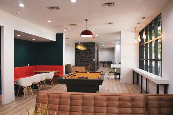 Modern communal room featuring a pool table, cozy seating with vibrant red cushions, stylish pendant lighting, and large windows for ample natural light.