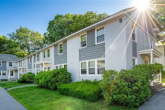 Charming apartment building surrounded by lush greenery and a sunlit path, offering a serene living environment.