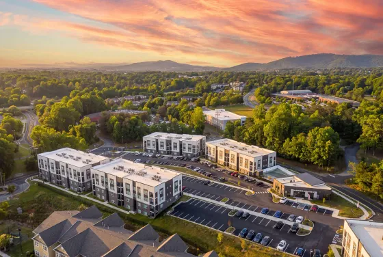 Aerial view of modern apartment complex surrounded by lush greenery at sunset, featuring ample parking and scenic mountain backdrop.